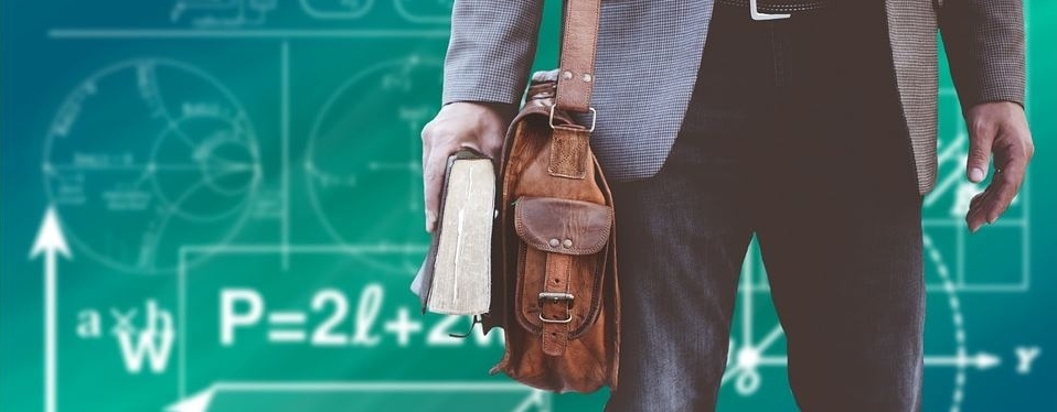 Bildausschnitt von Mann mit hellblauem Sacko und schwarzer Jeans, Hüfte bis Knie. Rechts trägt er ein braune Aktentasche aus Leder und hält ein dickes Buch in der Hand. Im Hintergrund ist eine grüne Tafel mit angedeuteten Formeln, Gleichungen und Skizzen zu sehen.