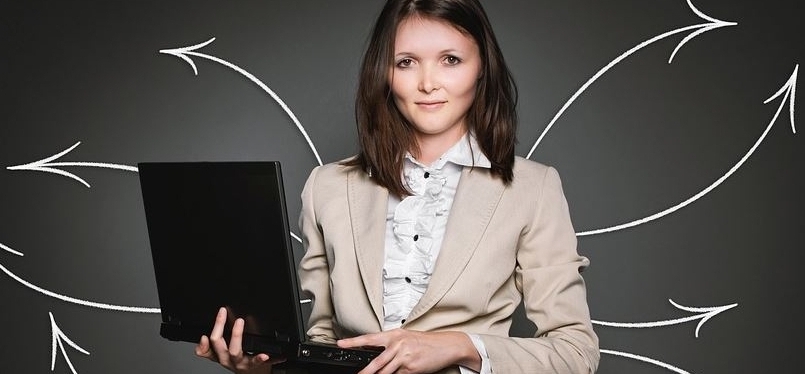 Geschäftsfrau mit dunklen, langen Haaren und einem aufgeklappten Laptop in der Hand. Im Hintergrund sind auf dunklem Grund weiße Kreidepfeile in verschiedene Richtungen kreisförmig angeordnet.