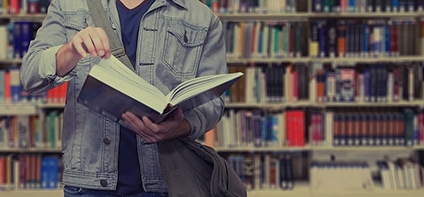 Ausschnitt lesender Student mit aufgeschlagenem Buch in der Hand. Im Hintergrund Bücherregale einer Bibliothek.