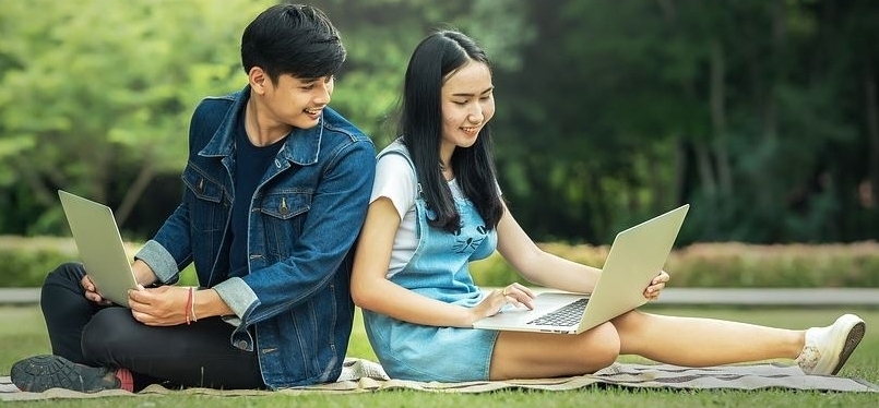 Zwei asiatische junge Menschen sitzen auf einer Decke im Park, den Laptop auf dem Schoß. Mann sitzt links, Frau rechts.