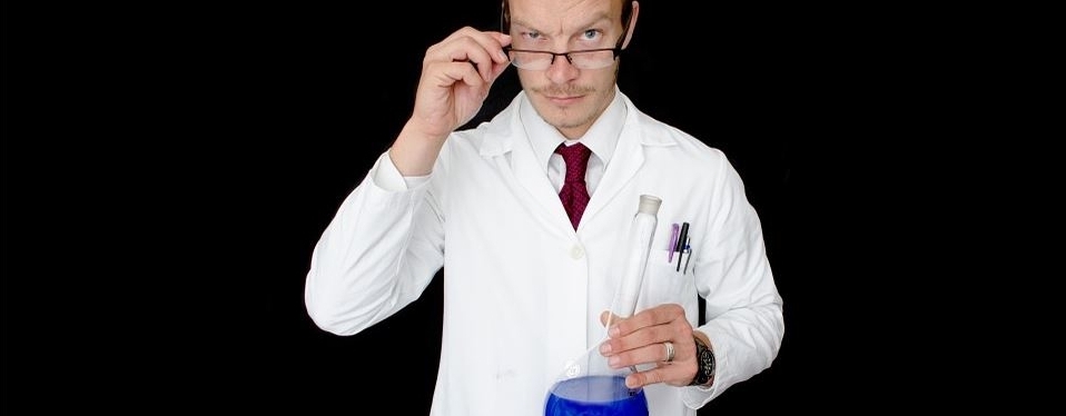 Laborant hält Kolbengefäß mit blauer Flüßigkeit in linker Hand und zückt die Brille auf der Nase nach vorn mit der rechten Hand.