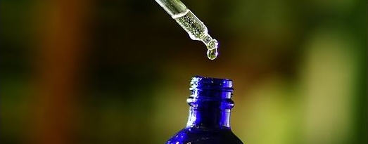 Kleine, dunkelblaue Glasflasche, ähnlich Nasentropfen, über der eine Pipette mit durchsichtiger Flüssigkeit zu sehen ist.