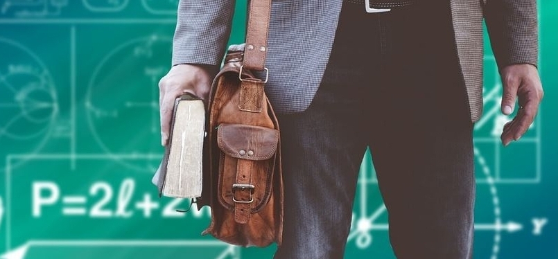 Bildausschnitt von Mann mit hellblauem Sacko und schwarzer Jeans, Hüfte bis Knie. Rechts trägt er ein braune Aktentasche aus Leder und hält ein dickes Buch in der Hand. Im Hintergrund ist eine grüne Tafel mit angedeuteten Formeln, Gleichungen und Skizzen zu sehen.